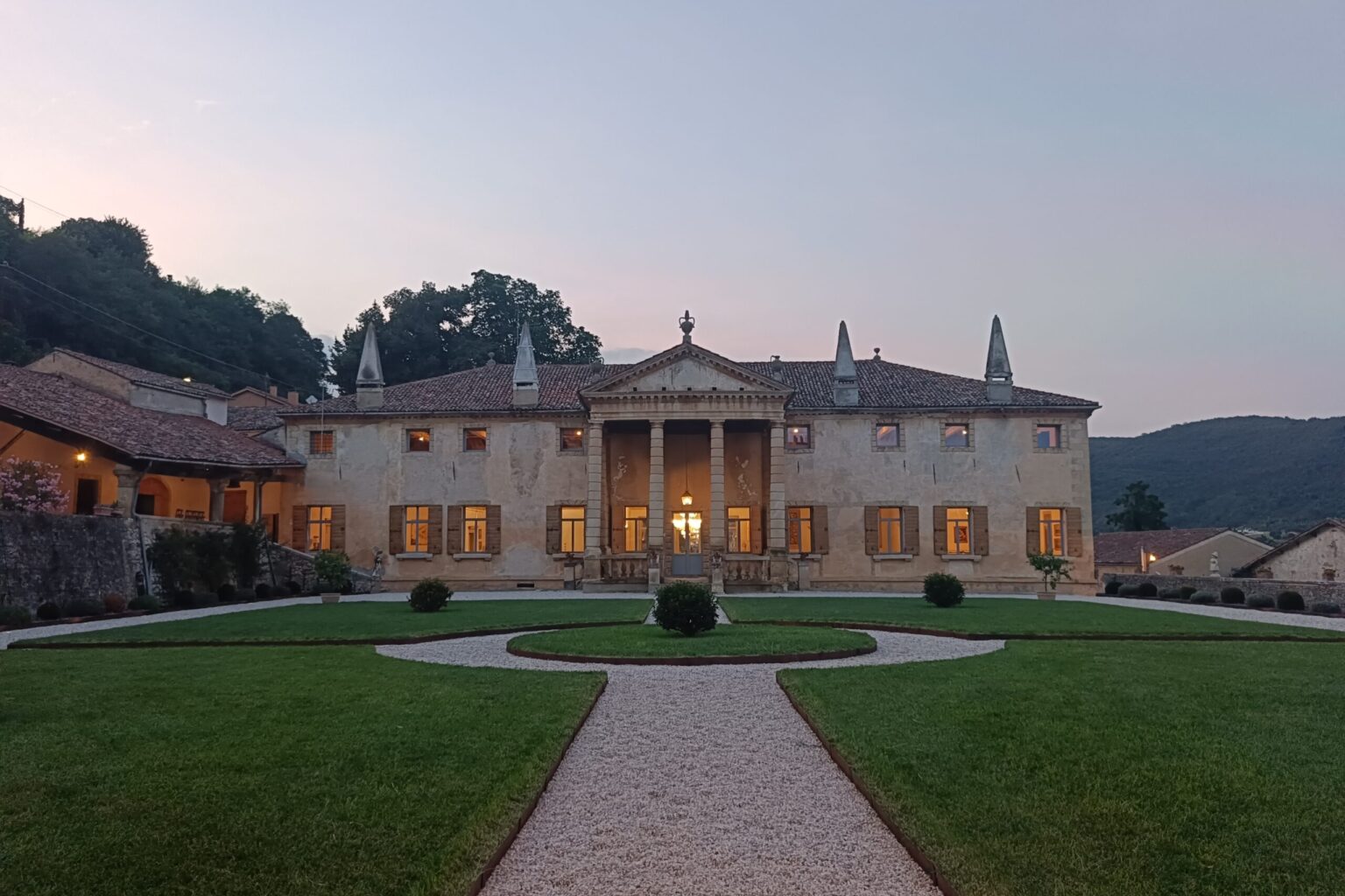 Elegant historic villa with a grand entrance, surrounded by manicured gardens and scenic hills at dusk. The building features classical architecture with tall columns, intricate details, and large windows that illuminate the exterior. | Only on SopranoVillas