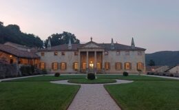 Elegant historic villa with a grand entrance, surrounded by manicured gardens and scenic hills at dusk. The building features classical architecture with tall columns, intricate details, and large windows that illuminate the exterior.