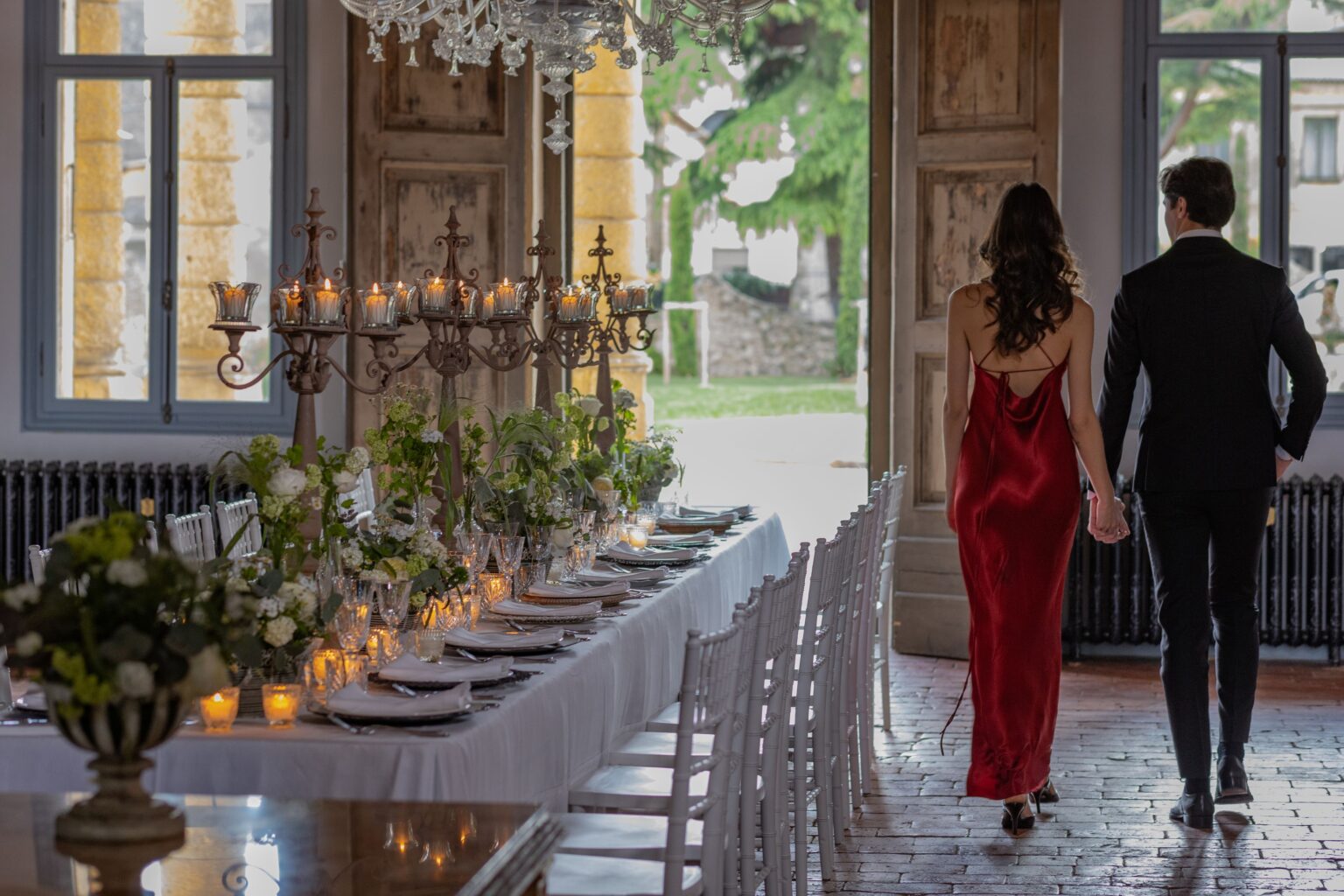 Couple walking hand in hand towards an elegantly set dining table adorned with candles and floral arrangements in a beautifully decorated venue, featuring large windows and a chandelier. | Only on SopranoVillas