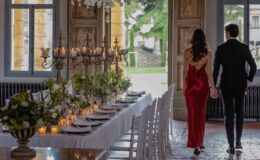 Couple walking hand in hand towards an elegantly set dining table adorned with candles and floral arrangements in a beautifully decorated venue, featuring large windows and a chandelier.