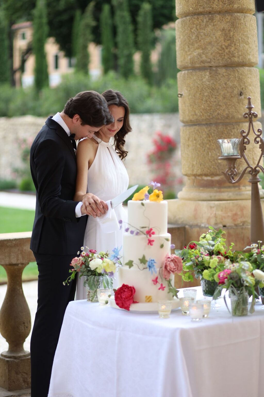 Couple in formal attire cutting a beautifully decorated wedding cake surrounded by floral arrangements and candles, set in an elegant outdoor venue. | Only on SopranoVillas