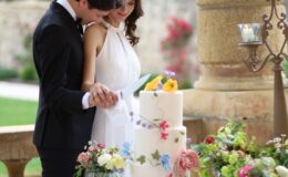 Couple in formal attire cutting a beautifully decorated wedding cake surrounded by floral arrangements and candles, set in an elegant outdoor venue.