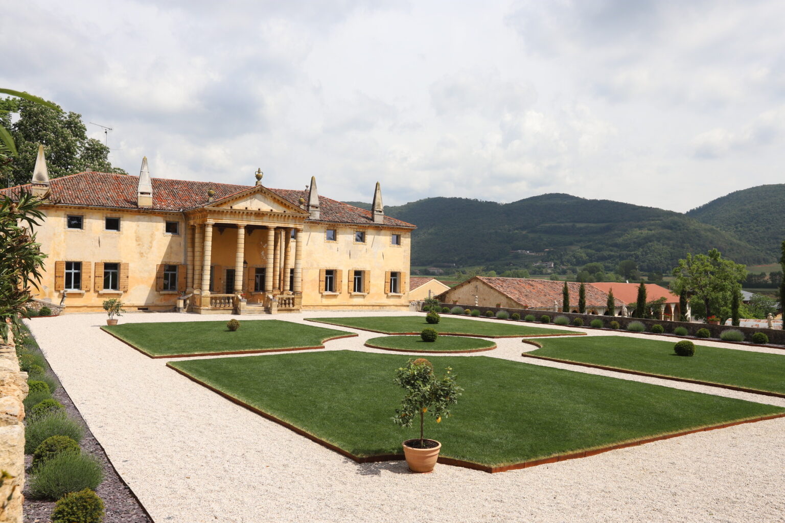 Elegant villa with classical architecture surrounded by meticulously landscaped gardens and rolling hills in the background. The property features a grand entrance with columns, manicured lawns, and decorative plants, showcasing a blend of luxury and natural beauty. Ideal for real estate listings or travel destinations. | Only on SopranoVillas
