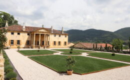 Elegant villa with classical architecture surrounded by meticulously landscaped gardens and rolling hills in the background. The property features a grand entrance with columns, manicured lawns, and decorative plants, showcasing a blend of luxury and natural beauty. Ideal for real estate listings or travel destinations.
