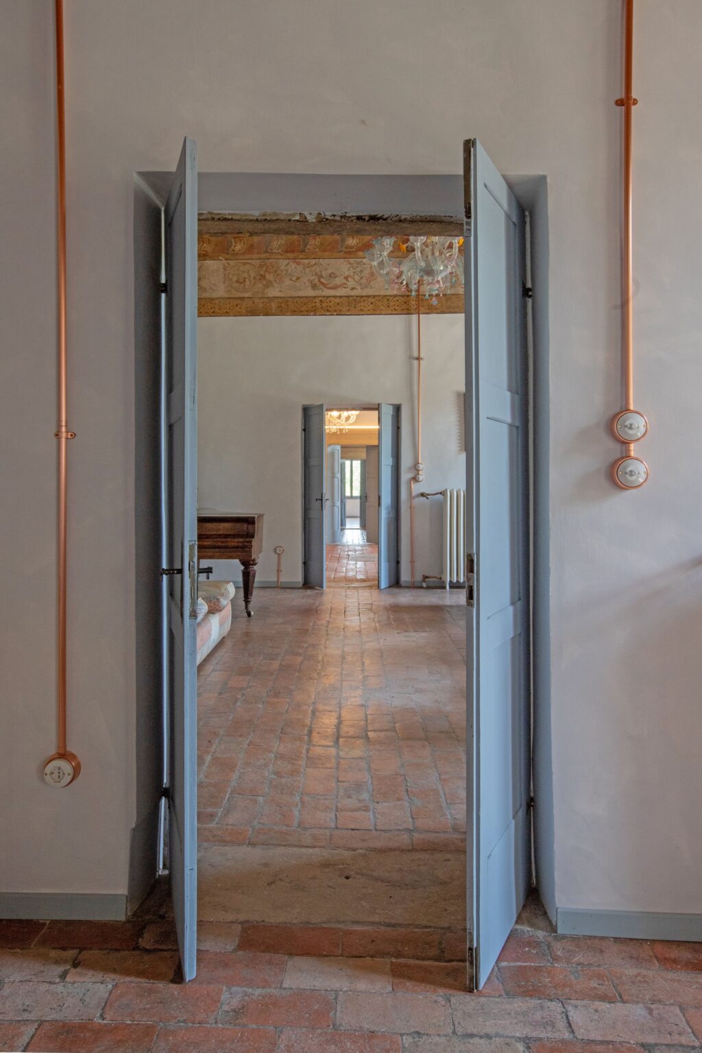 Interior view of a stylish room featuring open blue doors, exposed copper piping, and terracotta flooring. The space showcases a vintage wooden piano and a glimpse of a chandelier in the background, highlighting a blend of modern and classic design elements. | Only on SopranoVillas