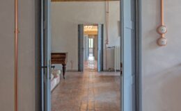 Interior view of a stylish room featuring open blue doors, exposed copper piping, and terracotta flooring. The space showcases a vintage wooden piano and a glimpse of a chandelier in the background, highlighting a blend of modern and classic design elements.