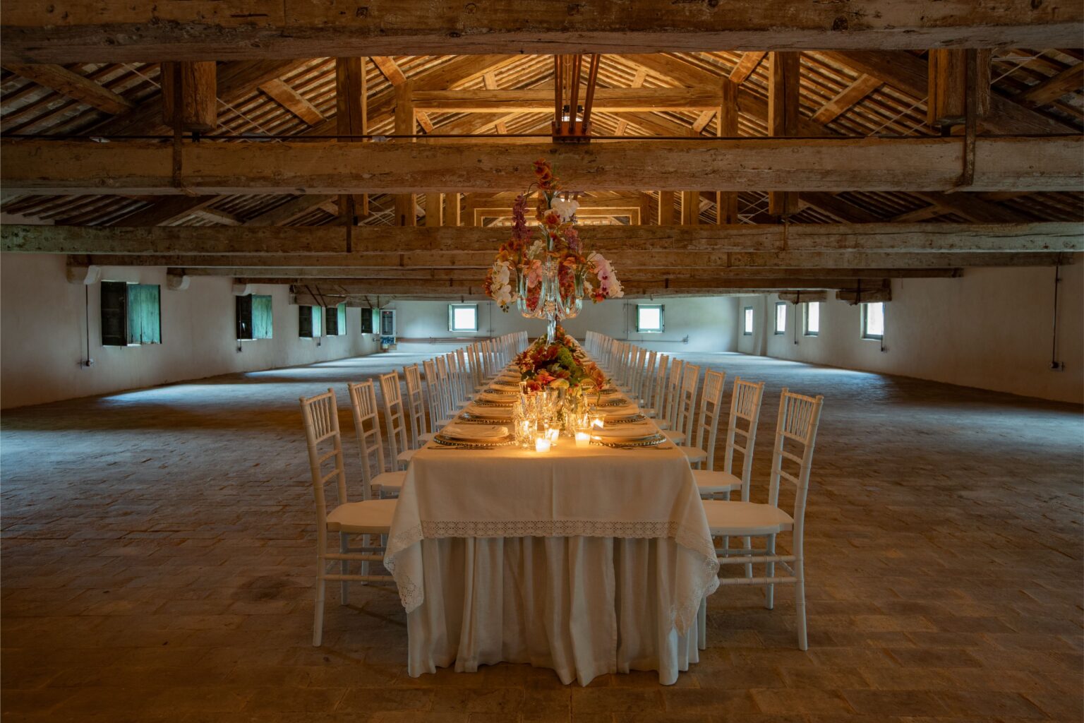 Elegant long dining table set for a formal event in a spacious rustic venue, featuring white chairs, floral centerpieces, and soft candlelight, under wooden beams and bright windows. | Only on SopranoVillas