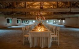 Elegant long dining table set for a formal event in a spacious rustic venue, featuring white chairs, floral centerpieces, and soft candlelight, under wooden beams and bright windows.