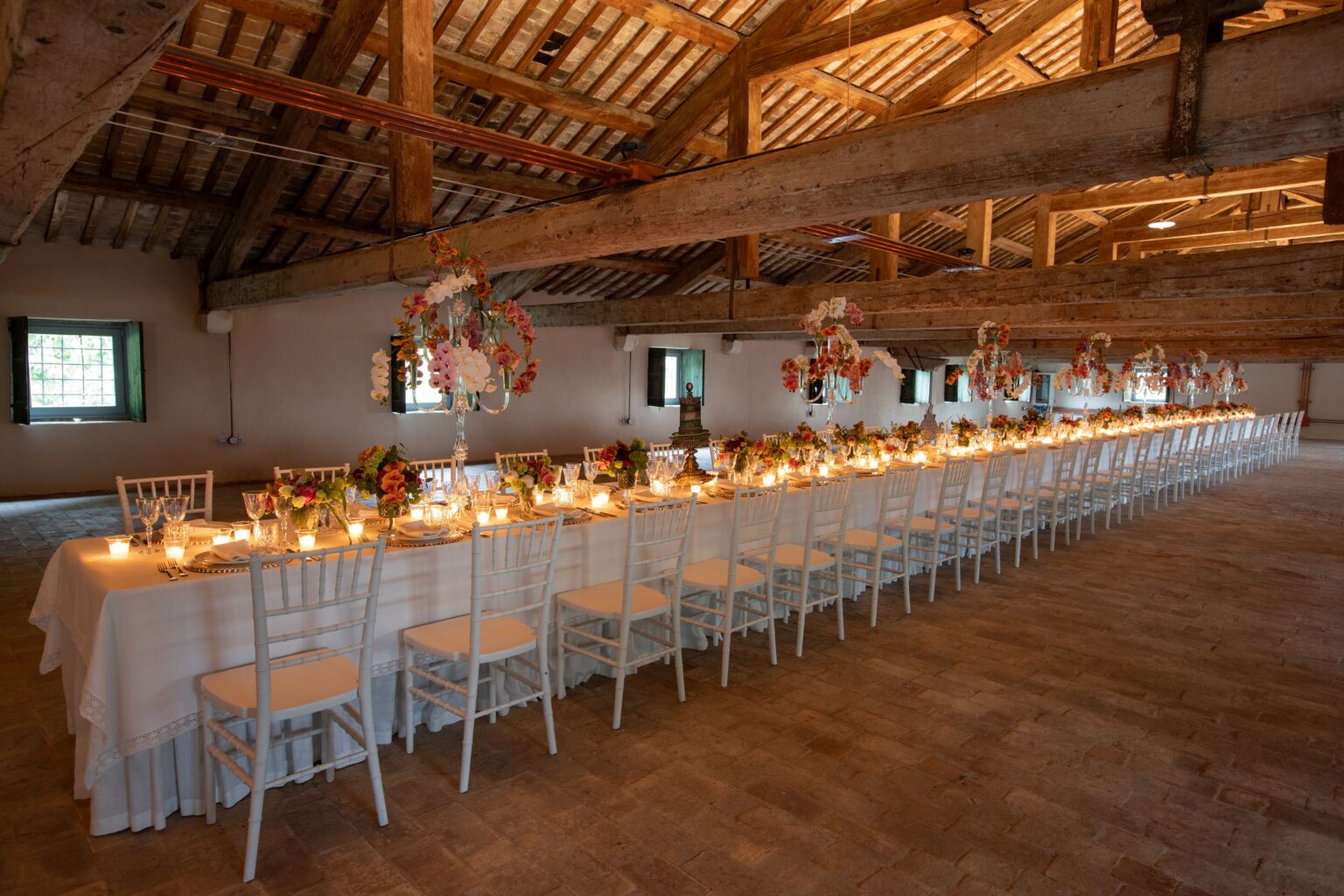 Elegant long dining table set for an event in a rustic venue, featuring white chairs, floral centerpieces, and soft candlelight, with exposed wooden beams and natural lighting. | Only on SopranoVillas
