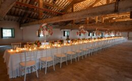 Elegant long dining table set for an event in a rustic venue, featuring white chairs, floral centerpieces, and soft candlelight, with exposed wooden beams and natural lighting.