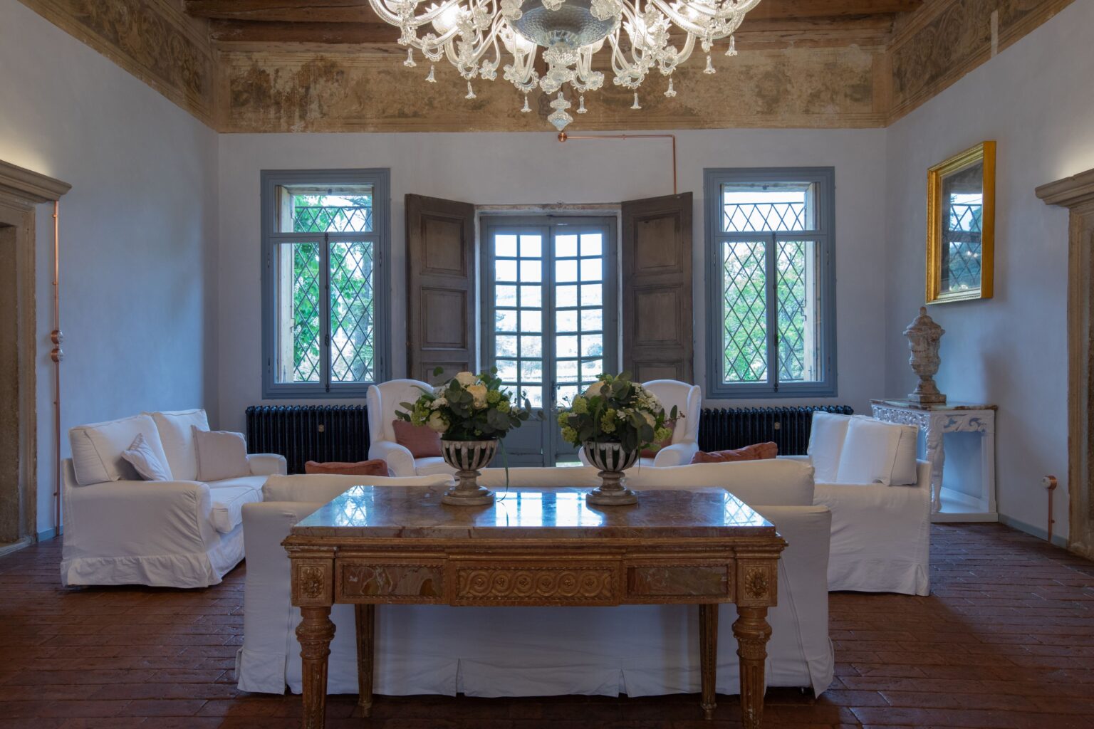 Elegant living room with white upholstered sofas, a decorative wooden table, and a vintage chandelier, featuring large windows that provide natural light and views of greenery. The space showcases classic decor elements, including floral arrangements and a golden-framed mirror. | Only on SopranoVillas