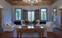 Elegant living room with white upholstered sofas, a decorative wooden table, and a vintage chandelier, featuring large windows that provide natural light and views of greenery. The space showcases classic decor elements, including floral arrangements and a golden-framed mirror.