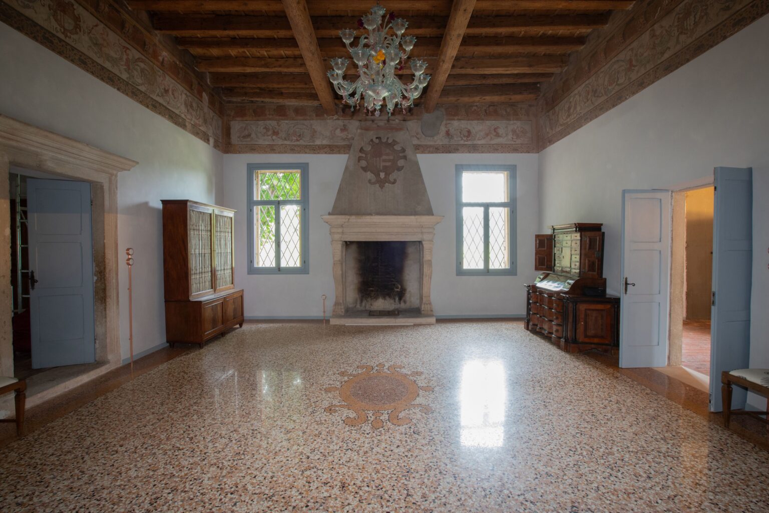 Elegant interior of a historic living room featuring a decorative chandelier, wooden beams, and a stone fireplace. The space showcases antique furniture, large windows with diamond-patterned glass, and a beautifully patterned terrazzo floor. | Only on SopranoVillas