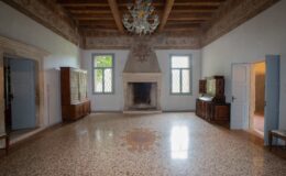 Elegant interior of a historic living room featuring a decorative chandelier, wooden beams, and a stone fireplace. The space showcases antique furniture, large windows with diamond-patterned glass, and a beautifully patterned terrazzo floor.