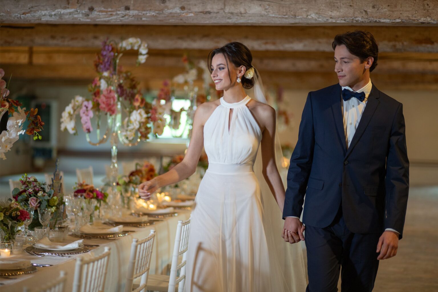 Bride and groom walking hand in hand in a beautifully decorated wedding venue, featuring elegant floral arrangements, crystal candelabras, and a long dining table set for a celebration. The bride wears a flowing white gown, while the groom is dressed in a classic navy suit and bow tie. | Only on SopranoVillas