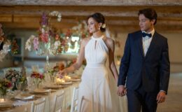 Bride and groom walking hand in hand in a beautifully decorated wedding venue, featuring elegant floral arrangements, crystal candelabras, and a long dining table set for a celebration. The bride wears a flowing white gown, while the groom is dressed in a classic navy suit and bow tie.