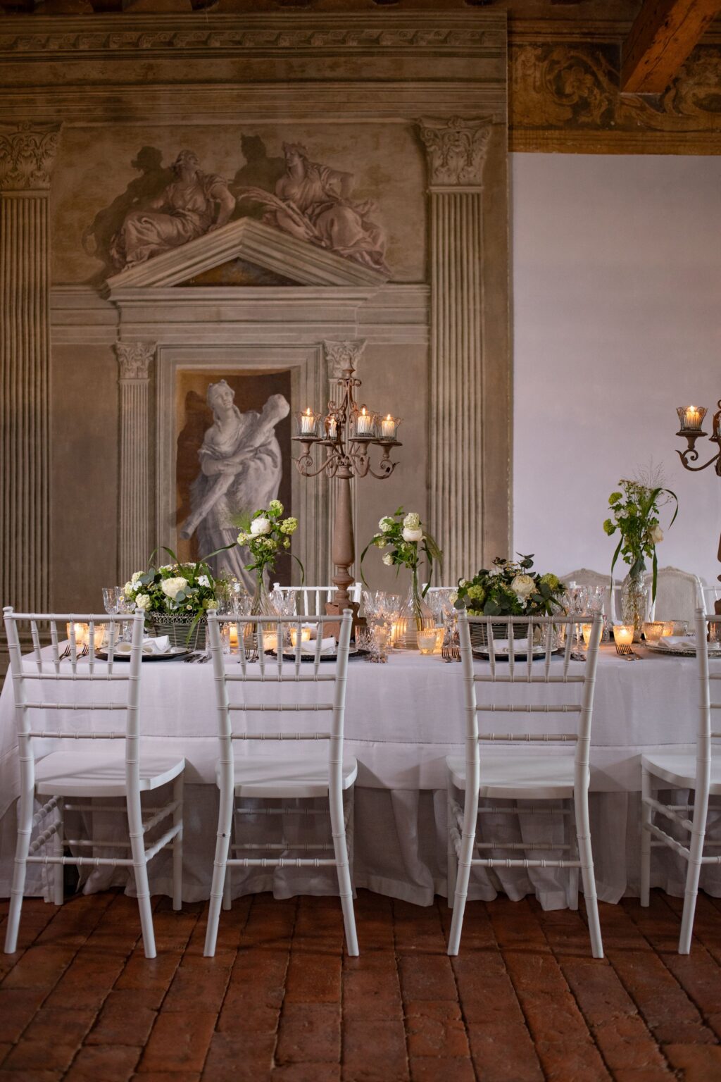 Elegant dining setup featuring a beautifully arranged table with white chairs, floral centerpieces, and candlelight, set against a backdrop of classical artwork and architectural details. Perfect for weddings or special events. | Only on SopranoVillas