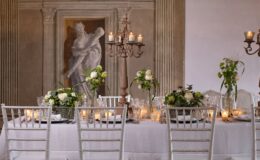 Elegant dining setup featuring a beautifully arranged table with white chairs, floral centerpieces, and candlelight, set against a backdrop of classical artwork and architectural details. Perfect for weddings or special events.