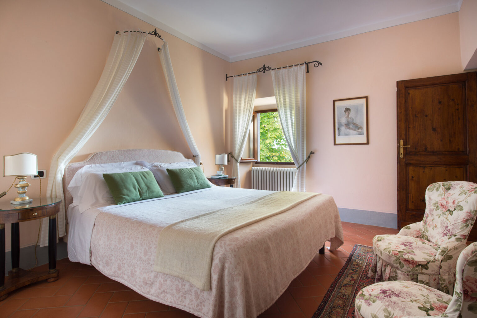 Cozy bedroom featuring a queen-sized bed with green pillows, elegant curtains, and floral armchairs. The room has warm peach walls, a wooden door, and a window with a view of greenery, creating a serene and inviting atmosphere. Perfect for a relaxing getaway. | Only on SopranoVillas