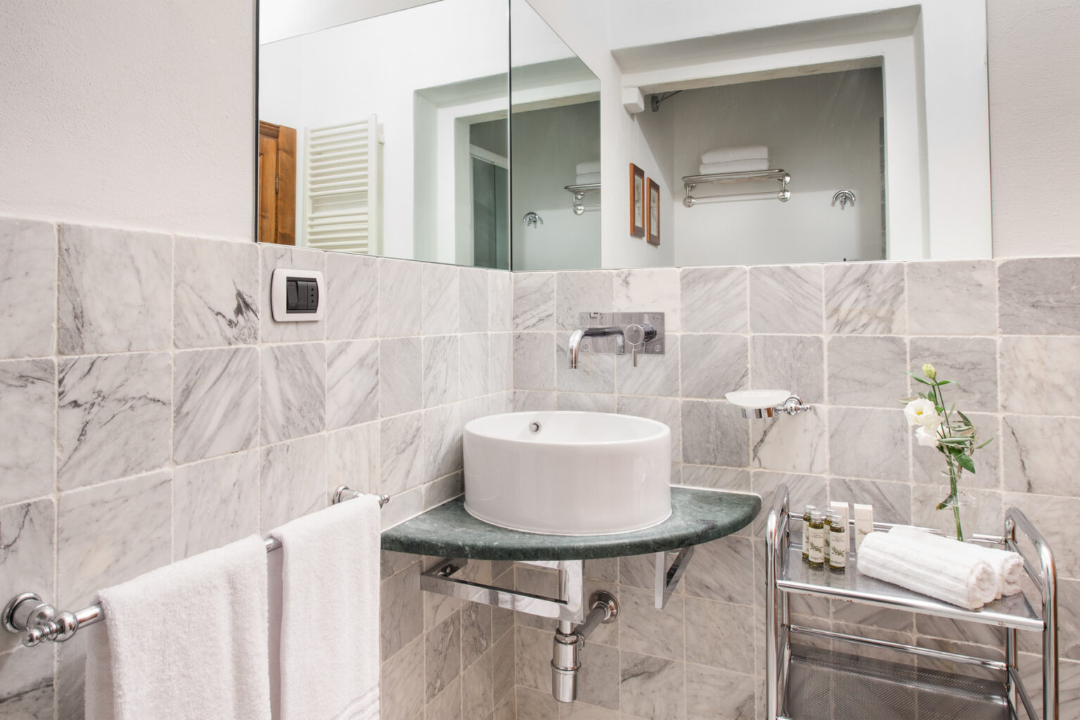 Modern bathroom featuring a marble tiled wall, a circular white sink on a green countertop, and a mirror. The space includes neatly arranged toiletries, fresh flowers, and plush towels, creating a stylish and inviting atmosphere. | Only on SopranoVillas