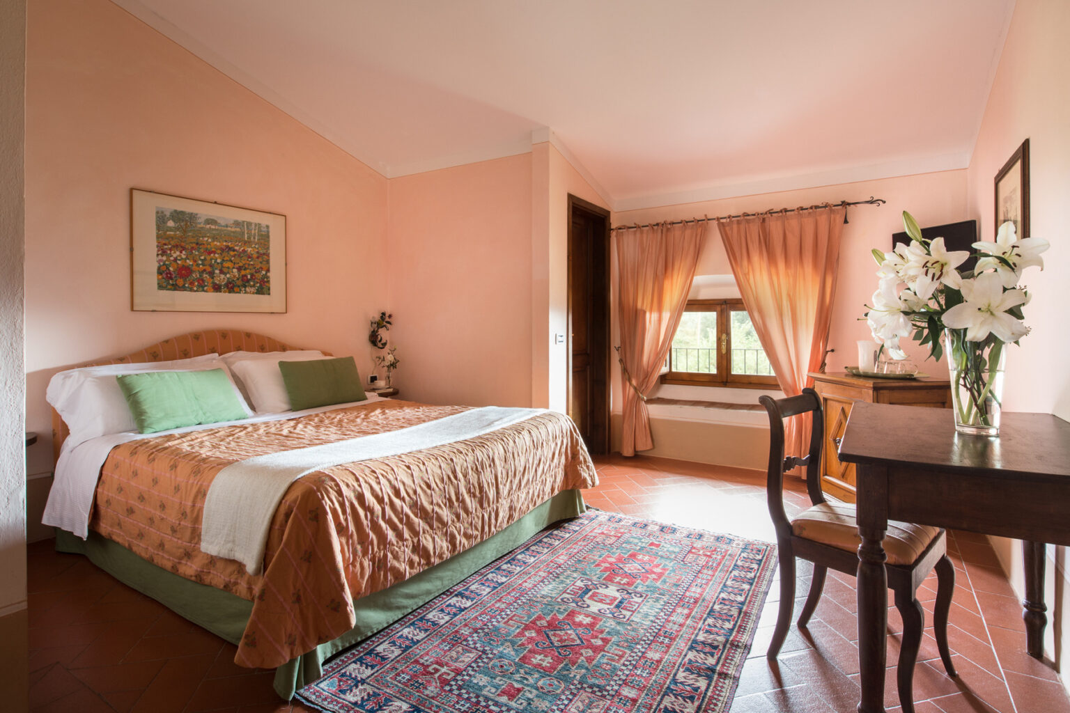 Cozy and elegantly decorated hotel room featuring a queen-sized bed with warm tones, a wooden desk, and a vibrant area rug. Natural light streams through large windows adorned with soft curtains, creating a welcoming atmosphere. Ideal for travelers seeking comfort and style. | Only on SopranoVillas