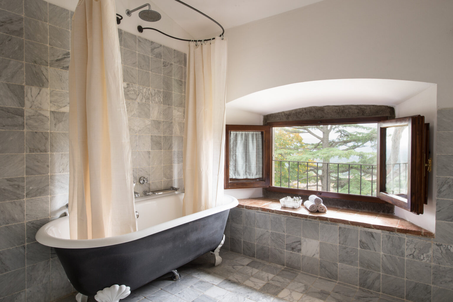 Elegant bathroom featuring a vintage clawfoot bathtub, light-colored curtains, and a window with a scenic view. The walls are adorned with stylish gray tiles, enhancing the modern yet classic aesthetic. | Only on SopranoVillas