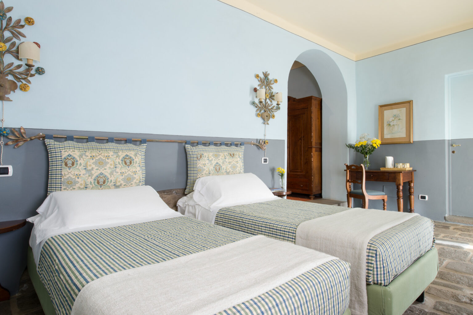 Cozy guest room featuring two twin beds with patterned bedding, light blue walls, and decorative wall art. A small wooden table with a vase of flowers adds charm to the space, complemented by natural light from the doorway. Perfect for a relaxing stay. | Only on SopranoVillas