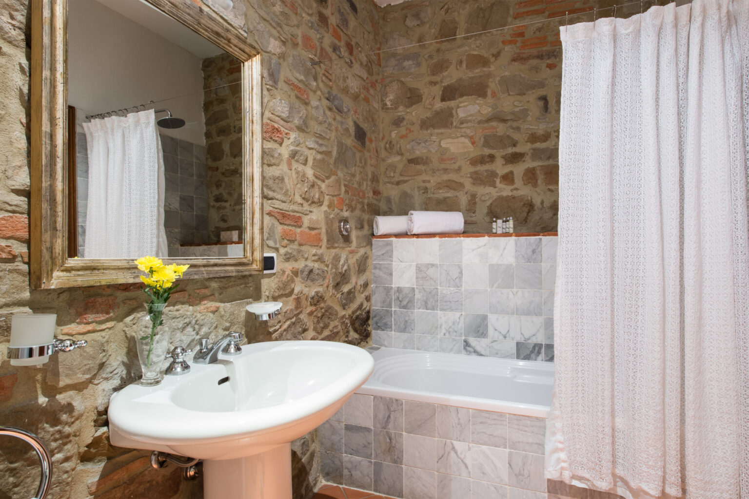 Modern bathroom with stone walls, featuring a white bathtub, sink, and a large mirror. Decorated with a small vase of yellow flowers and white shower curtain, creating a cozy and stylish atmosphere. | Only on SopranoVillas