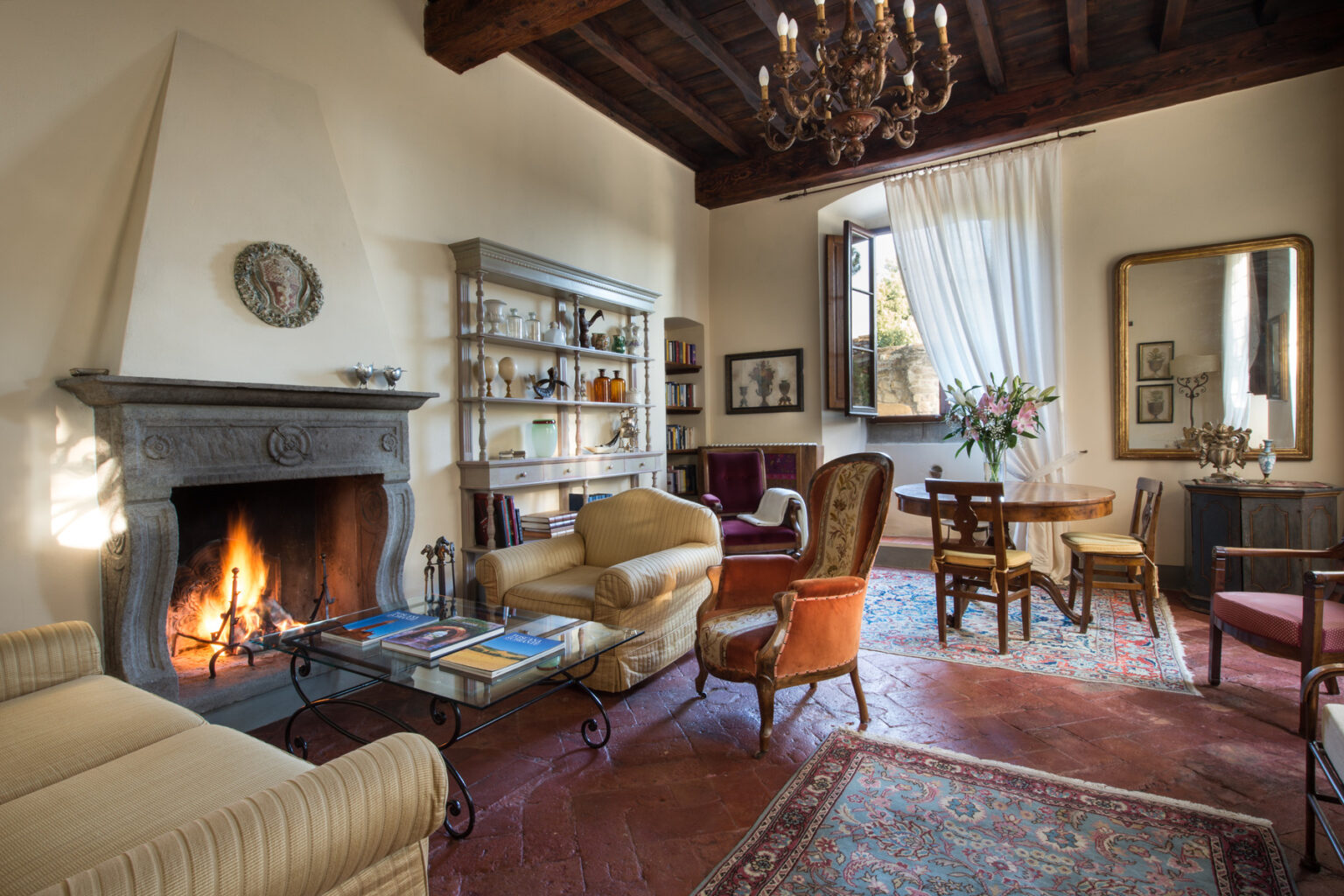 Cozy living room featuring a stone fireplace, elegant furnishings, and a chandelier, with natural light streaming through large windows. The space includes a comfortable sofa, armchairs, a dining table, and decorative shelves, creating a warm and inviting atmosphere. | Only on SopranoVillas