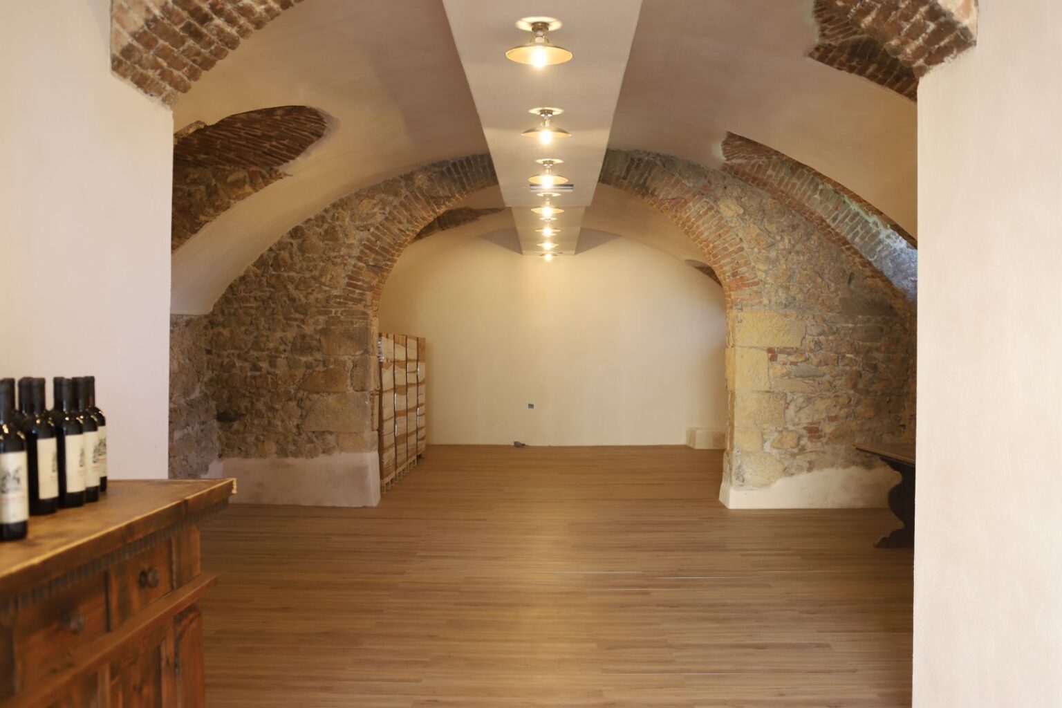 Interior view of a spacious wine cellar featuring exposed brick arches, wooden flooring, and shelves for wine storage, illuminated by modern ceiling lights. | Only on SopranoVillas