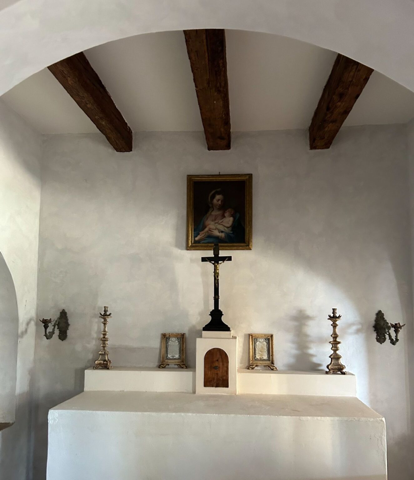 Interior view of a small chapel featuring a wooden beam ceiling, a framed painting of the Virgin Mary with child, a central crucifix, and decorative candlesticks on either side of the altar, all set against a textured white wall. | Only on SopranoVillas