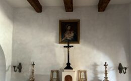 Interior view of a small chapel featuring a wooden beam ceiling, a framed painting of the Virgin Mary with child, a central crucifix, and decorative candlesticks on either side of the altar, all set against a textured white wall.