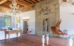 Elegant interior of a historic room featuring wooden beams, a crystal chandelier, and a blend of classical and modern decor. The room includes a decorative wall painting, an ornate wooden table, and a unique sculpture made from wood, all set against terracotta flooring.