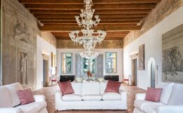 Elegant living room featuring white sofas with pink cushions, a stunning crystal chandelier, and intricately designed wall murals. Large windows provide natural light, showcasing a blend of classic and contemporary decor in a spacious, inviting atmosphere.