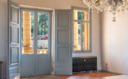 Elegant interior of a room featuring open double doors, a crystal chandelier, exposed wooden beams, and terracotta flooring. Natural light floods in, highlighting the decorative wall details and providing a view of a lush garden outside.