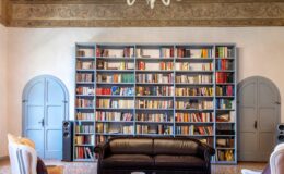 Elegant living room featuring a black leather sofa, two white armchairs, a glass coffee table, and a large bookshelf filled with colorful books. The room is illuminated by a stylish chandelier, with wooden beams on the ceiling and decorative wall details, creating a cozy and sophisticated atmosphere.