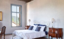 Cozy bedroom featuring a white wrought iron bed with navy blue and white bedding, antique wooden nightstands with lamps, and a large window offering scenic views. The room showcases rustic wooden beams and elegant decor, creating a serene atmosphere.