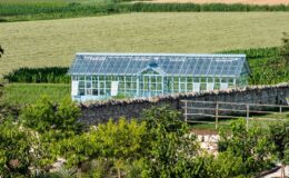 A picturesque view of a greenhouse surrounded by lush fields and trees, set against a backdrop of rolling hills and farmland. The greenhouse is made of glass, reflecting natural light, while the foreground features neatly arranged gardens with various plants and trees.