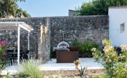 Outdoor patio featuring a rustic stone wall, a modern pizza oven, and a dining area with black furniture, surrounded by lush greenery and colorful flowers. Ideal for outdoor cooking and entertaining.