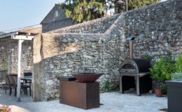 Outdoor kitchen area featuring a modern pizza oven and a fire pit, surrounded by a rustic stone wall and patio, ideal for entertaining and cooking.