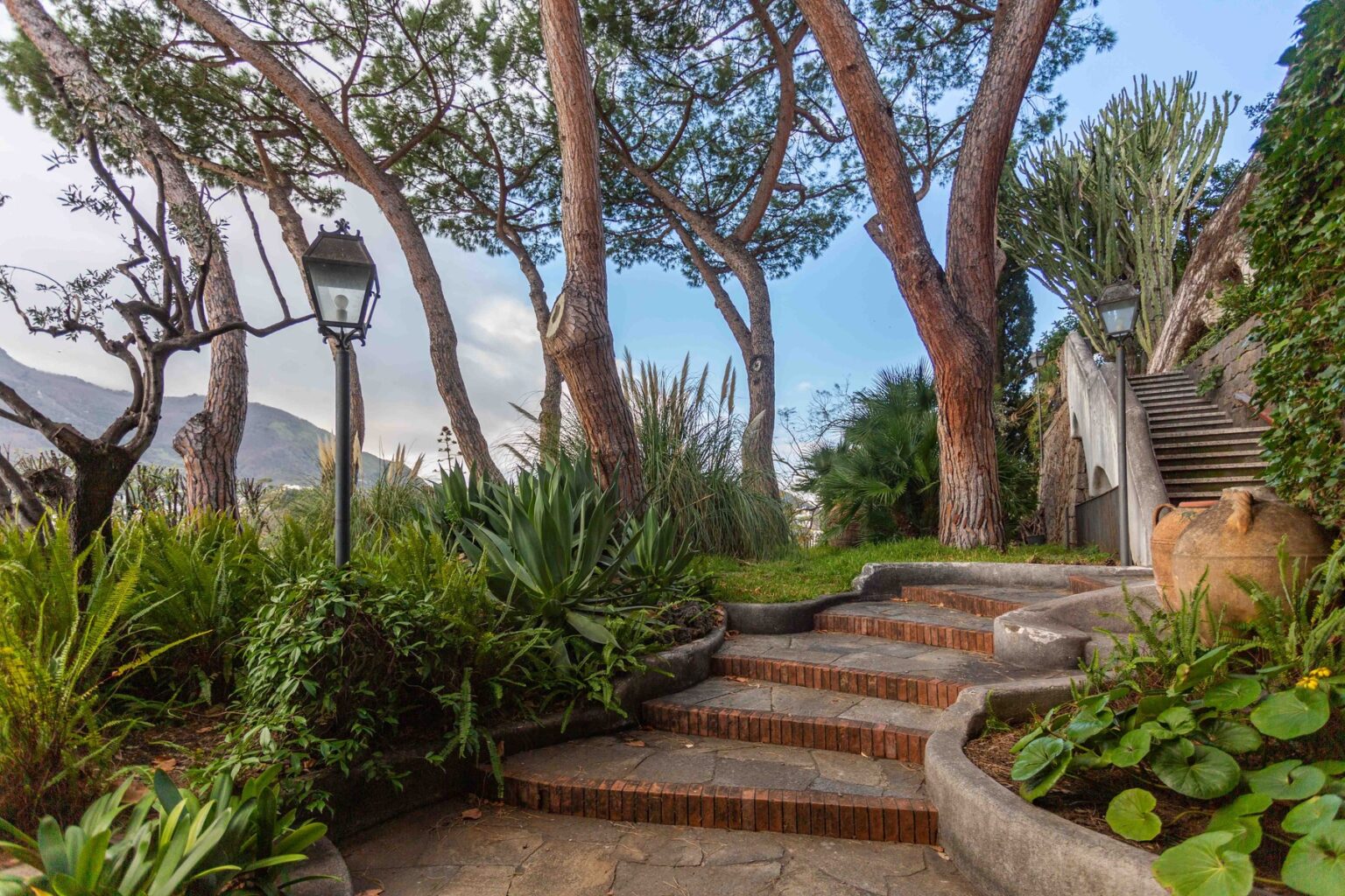 Lush garden pathway with stone steps, surrounded by tall trees and greenery, featuring decorative lanterns and a serene atmosphere. | Only on SopranoVillas