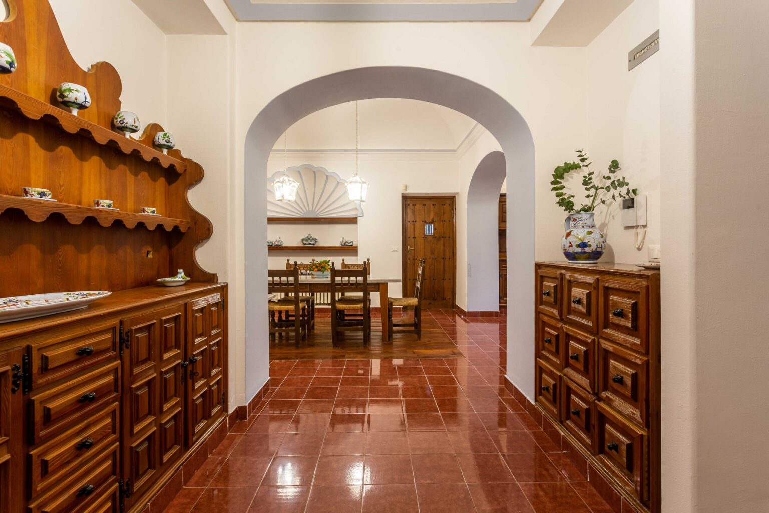 Interior view of a stylish dining area featuring wooden cabinets, a decorative shelf with ceramics, and a spacious archway leading to a well-lit dining table. The warm tones and polished floors create an inviting atmosphere. | Only on SopranoVillas