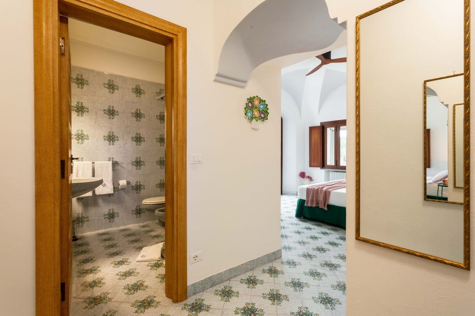 Interior view of a stylish bathroom entrance with decorative tiles, featuring a toilet and sink, leading to a well-lit bedroom with a green bed and wooden windows. A mirror reflects the elegant design, highlighting the spacious layout and modern decor. | Only on SopranoVillas