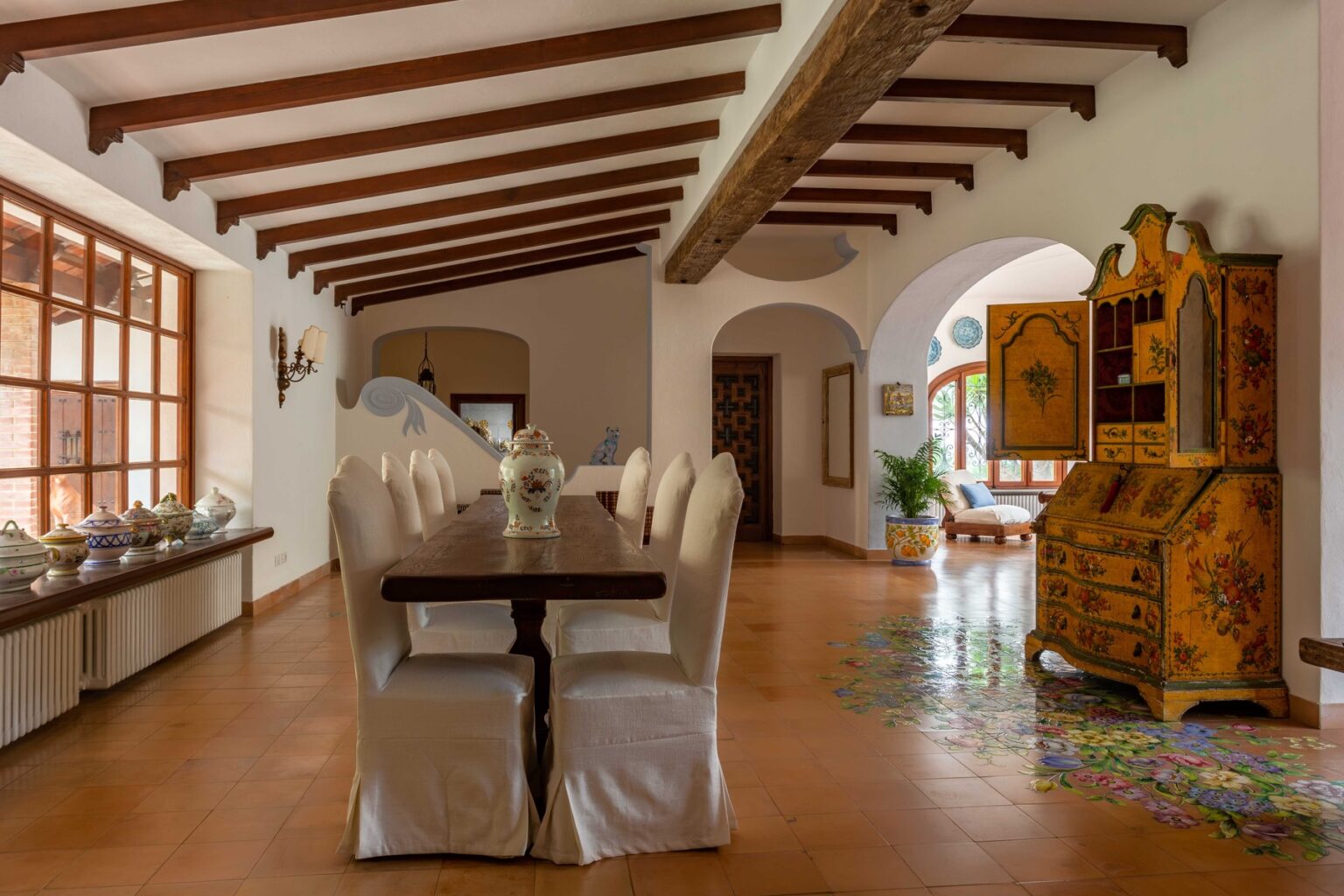 Spacious dining area featuring a long wooden table surrounded by white upholstered chairs, adorned with decorative pottery on the windowsill. The room showcases rustic wooden beams, an intricately painted yellow cabinet, and colorful floral designs on the floor, creating an inviting and stylish atmosphere. | Only on SopranoVillas