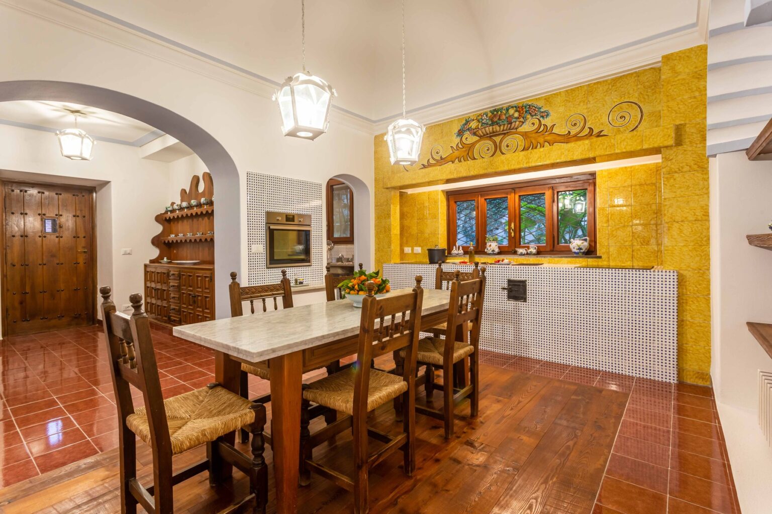 Cozy kitchen interior featuring a wooden dining table with six chairs, yellow accent wall, and rustic cabinetry. The space is illuminated by hanging lanterns and showcases a warm, inviting atmosphere perfect for family gatherings. | Only on SopranoVillas
