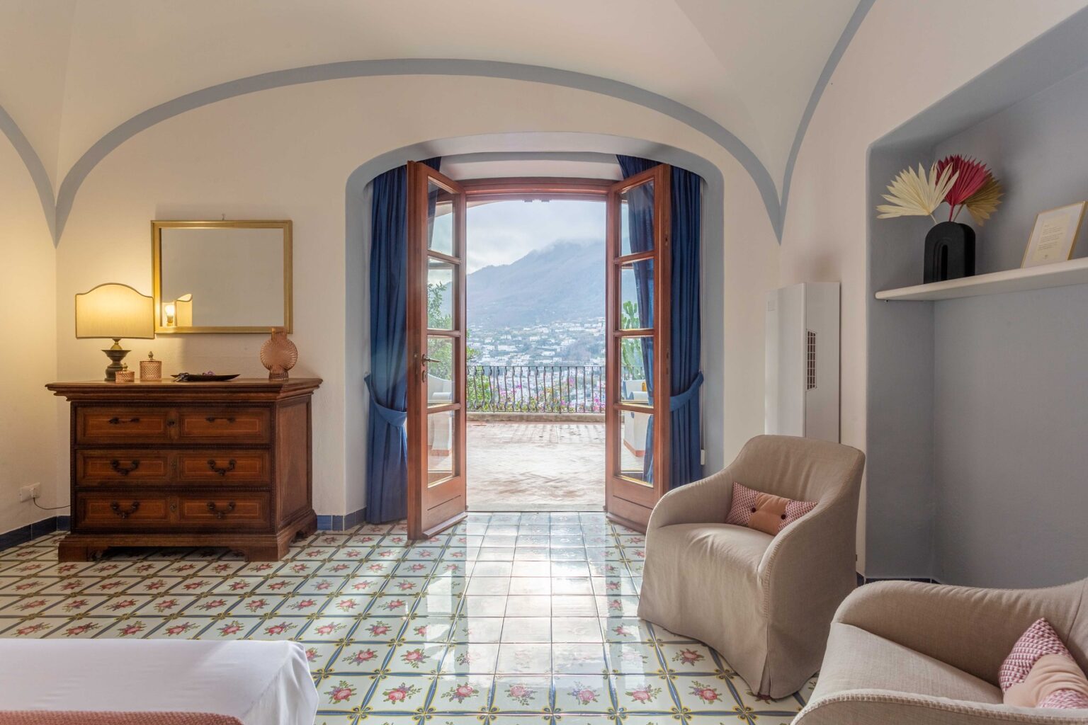 Cozy hotel room with a view, featuring a wooden dresser, comfortable chair, and open doors leading to a scenic balcony. The room showcases traditional tiled flooring and soft blue curtains, creating a serene atmosphere. | Only on SopranoVillas