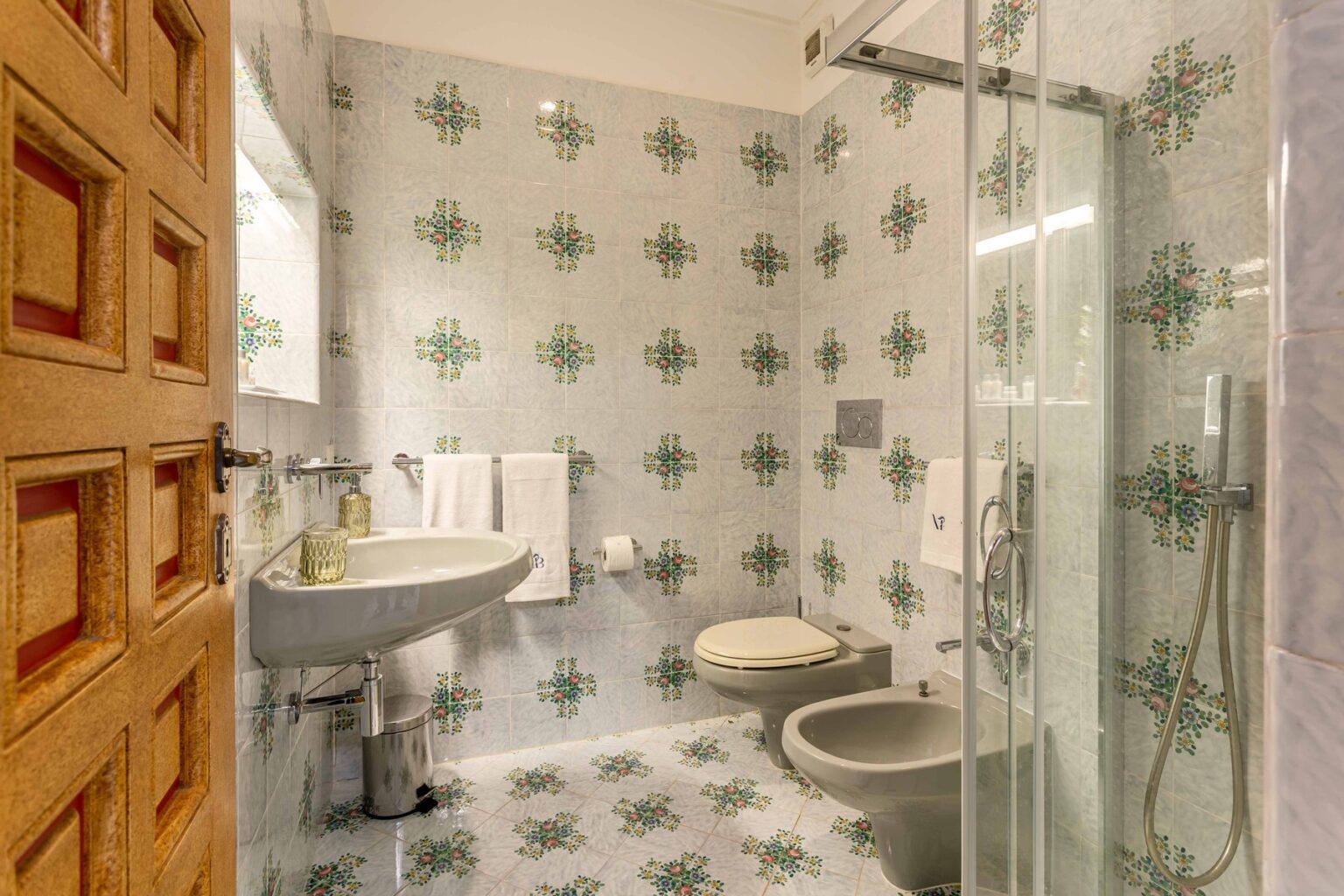 Modern bathroom featuring floral-patterned tiles, a glass shower enclosure, a pedestal sink, and a bidet. The space is well-lit and includes towels and decorative elements, showcasing a blend of functionality and aesthetic appeal. | Only on SopranoVillas