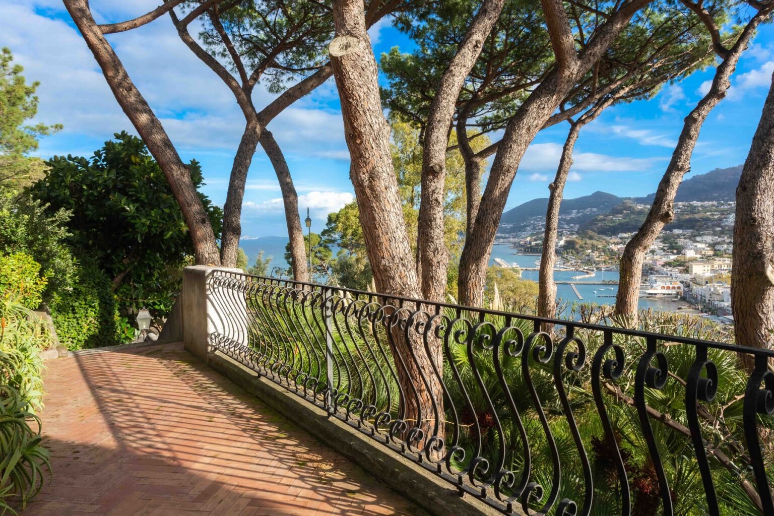 Scenic garden pathway with wrought iron railing surrounded by tall trees, overlooking a coastal view with mountains in the distance. | Only on SopranoVillas