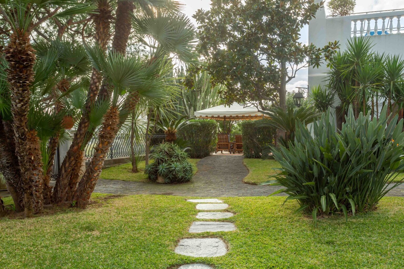Lush garden pathway leading to a shaded seating area, surrounded by tropical plants and trees, creating a serene outdoor retreat. | Only on SopranoVillas