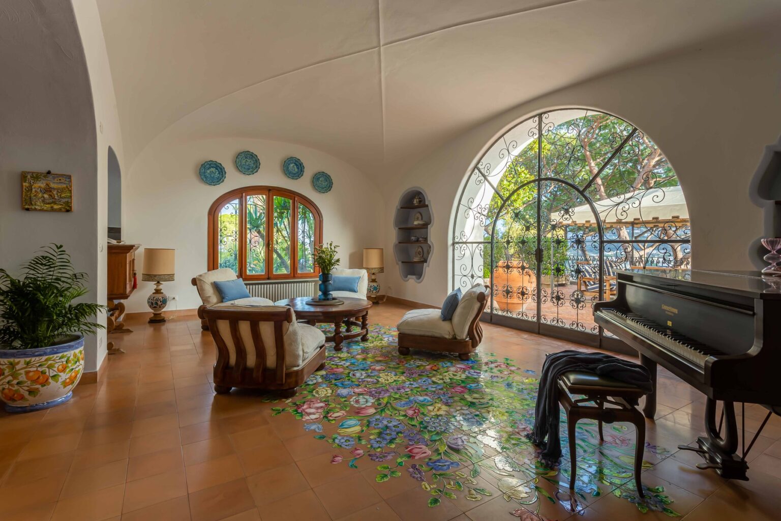 Bright and airy living room featuring a grand piano, comfortable seating, and large arched windows that provide natural light and a view of the garden. Colorful tile flooring adds a vibrant touch to the space. | Only on SopranoVillas